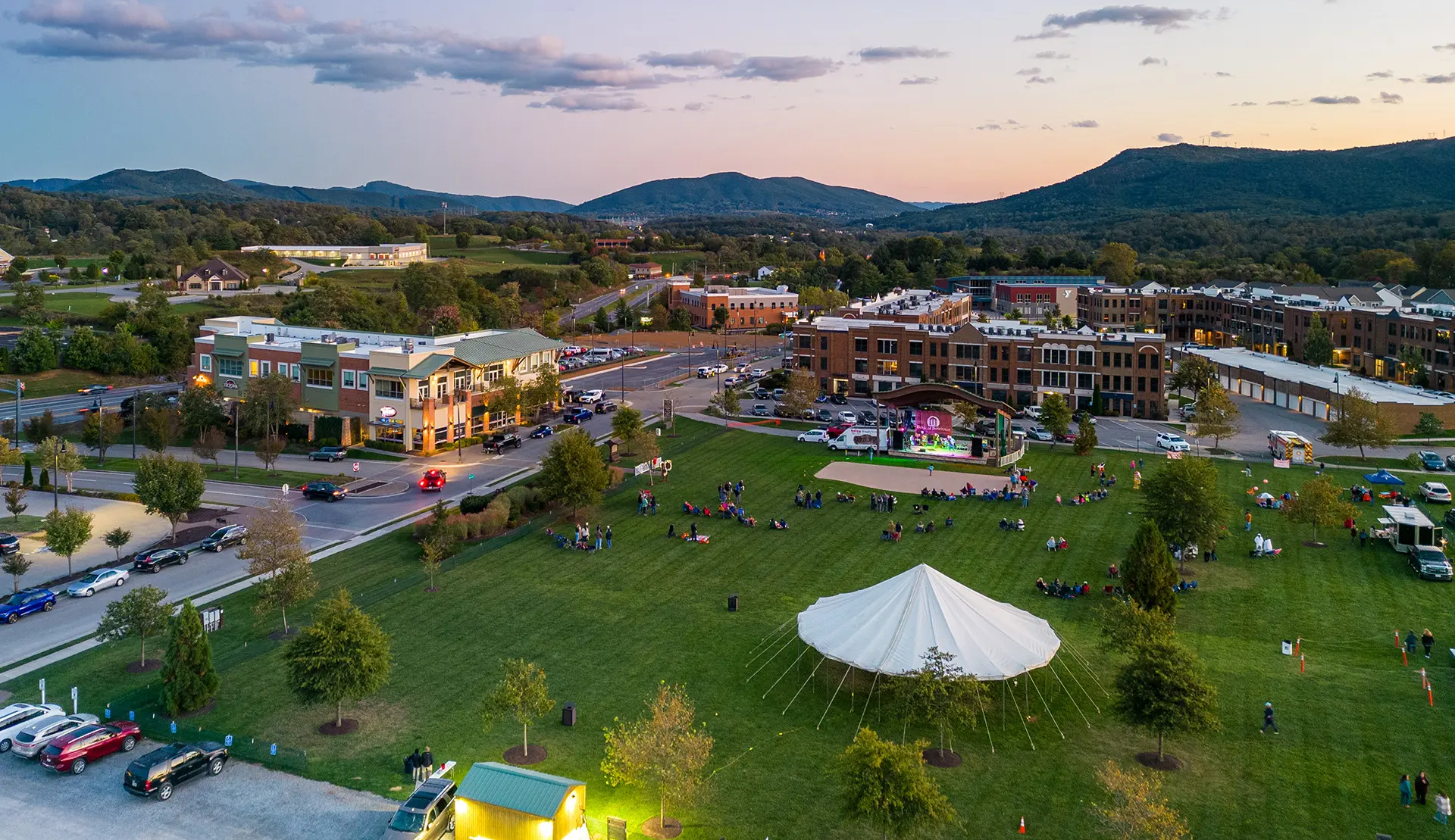 Aerial photo of the entire Daleville Town Center during sunset on a summer night. There are vibrant green lawns, apartment building, townhomes, and shops.