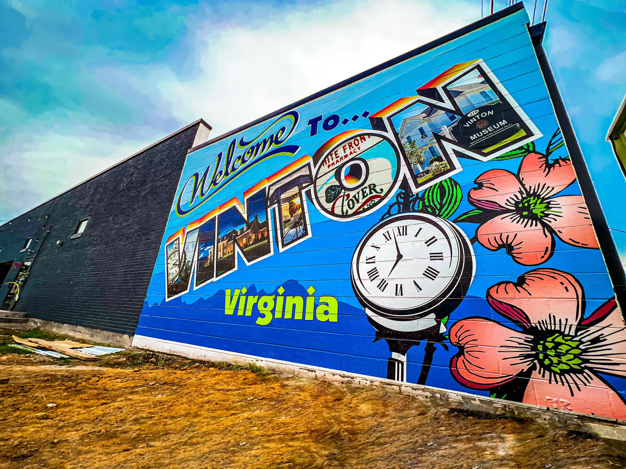 Hand=painted mural in downtown Vinton, VA. It reads "Welcome to Vinton Virginia" with blue mountains along the bottom, pink dogwood flowers, and the clock that is in downtown Vinton. Painted inside of the "VINTON" are different Vinton landmarks.