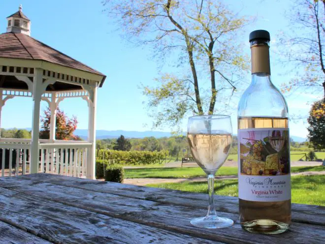 Glass of wine and a bottle of wine sitting on a picnic table at Virginia Mountain Winery. There is a gazebo and mountain view in the background.