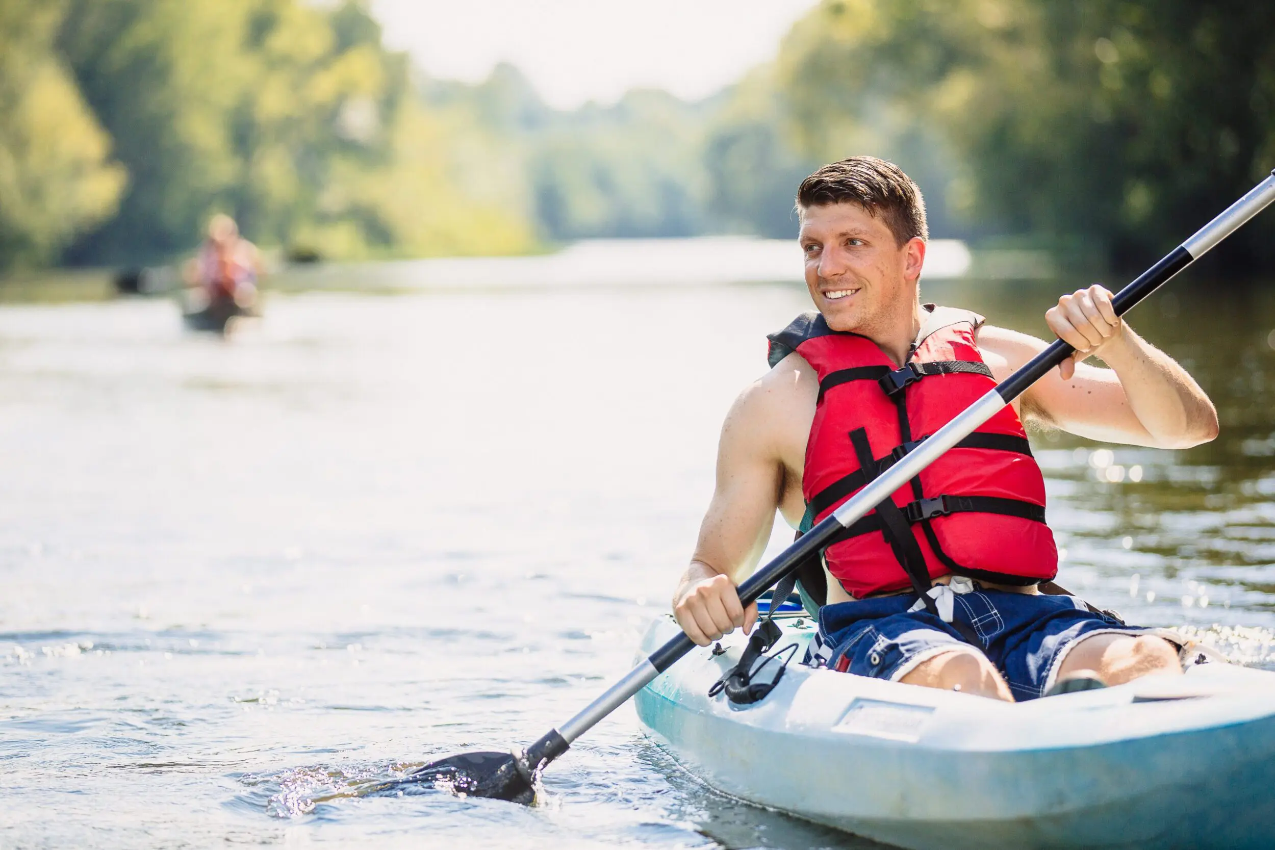 White man in his late 20s or early 30s smiles as he paddles in a blue sit-on-top kayak on the James River. He is wearing a read life jacket.