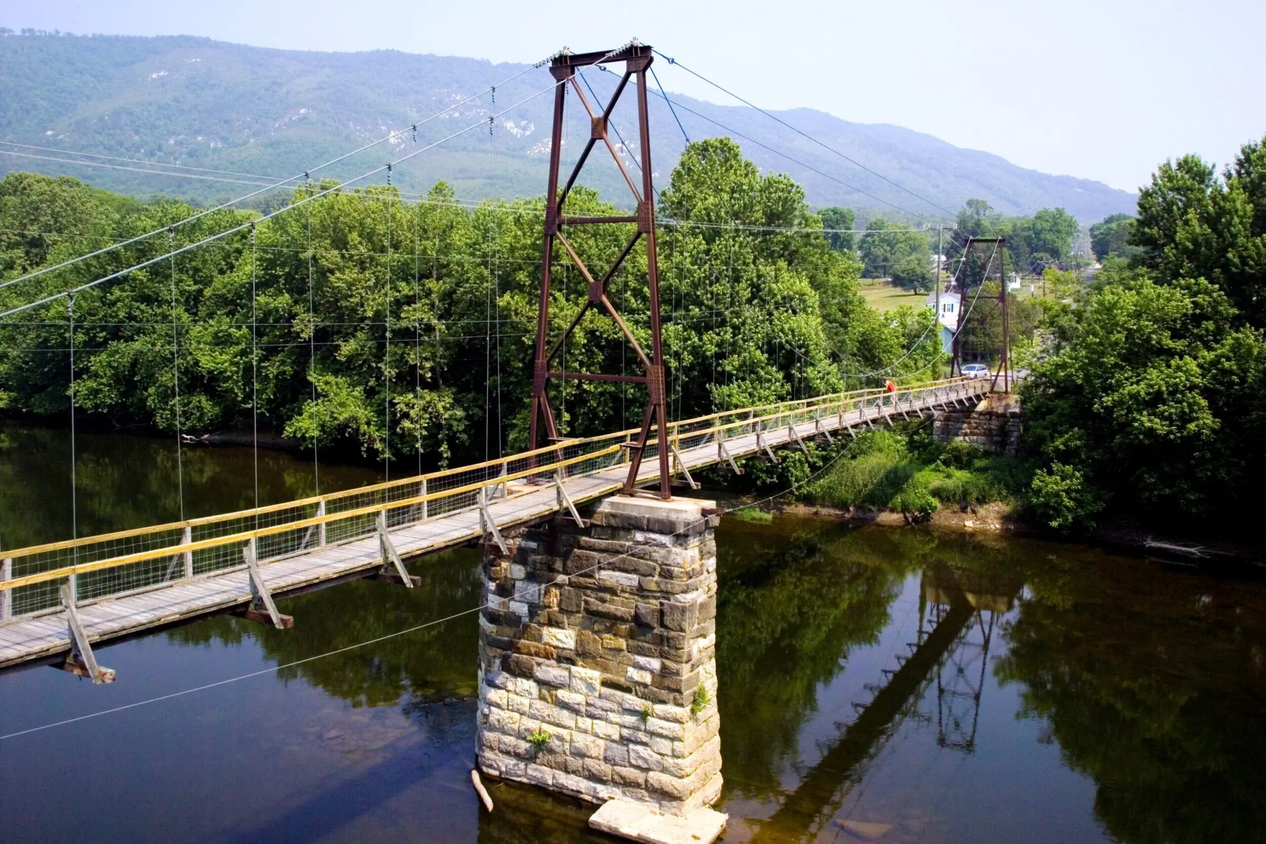 Swinging Bridge in Buchanan, VA. The James River is beneath the bridge, reflecting the bridge and woods that line the river with the mountains in the background.