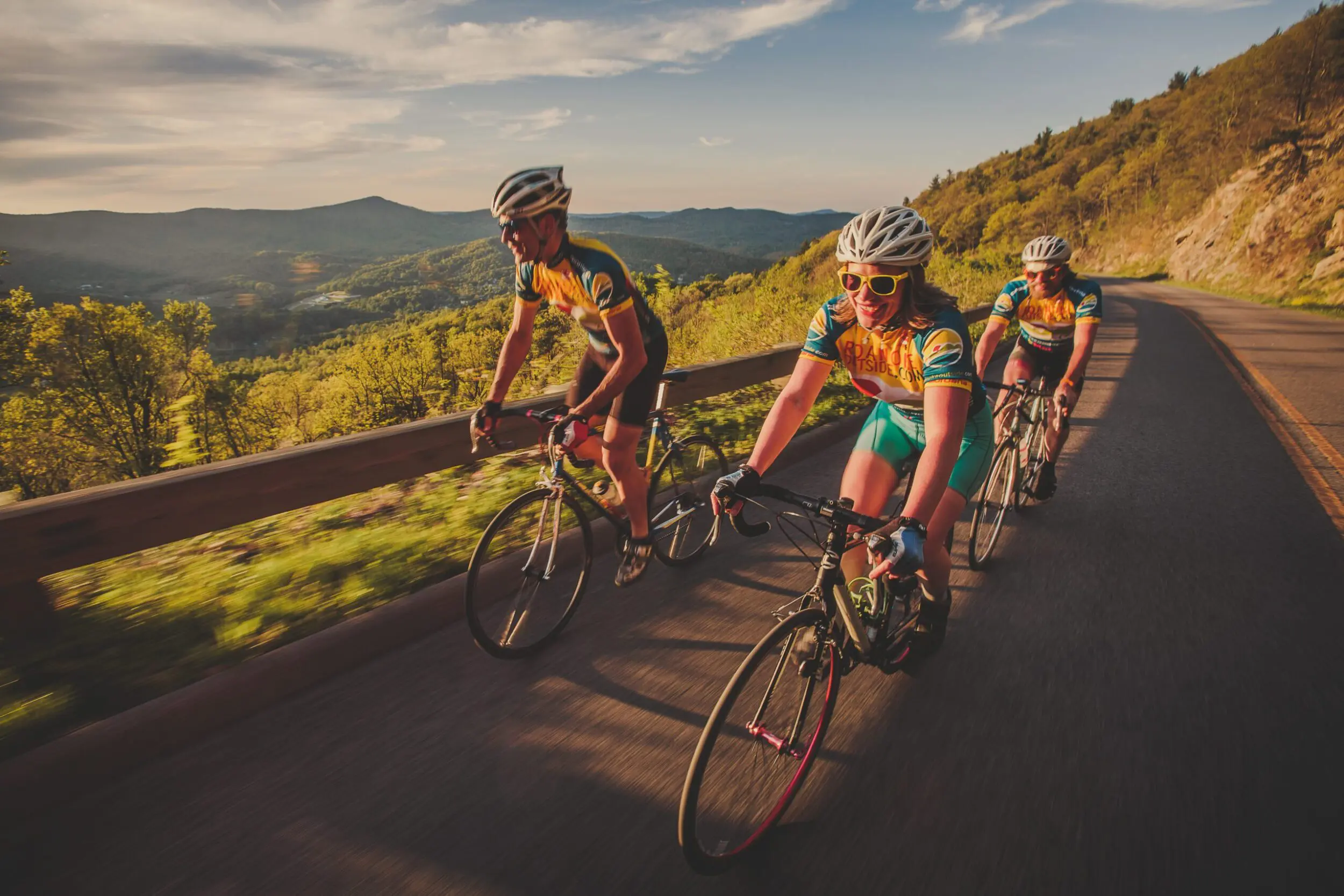 Three road cyclists in cycling kits bike up a mountain on the Blue Ridge Parkway at Golden Hour. They're all facing ahead and smiling as they pedal.