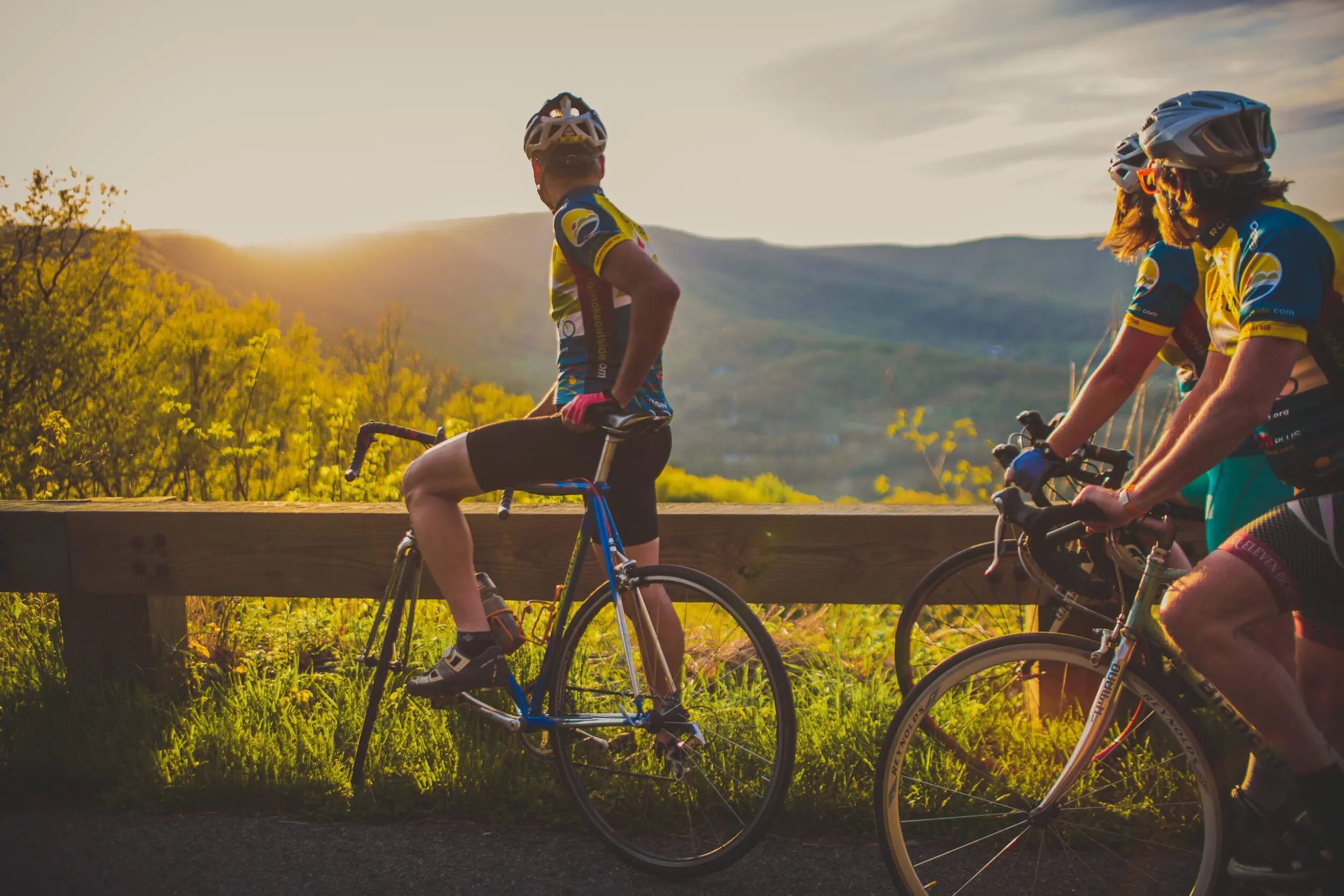 Three road cyclists in colorful cycling kits pause on the Blue Ridge Parkway as they look out over a beautiful vista of green mountains during sunset.