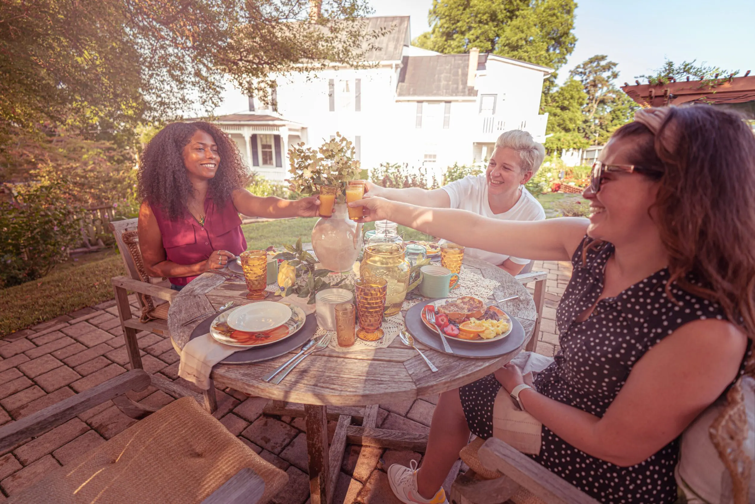 Three women, one black and two white, cheers one another at brunch at the Claiborne Bed and Breakfast in Rocky Mount, VA. There is tasty brunch foods on the plates in front of each woman, and there's a white colonial home in the background.