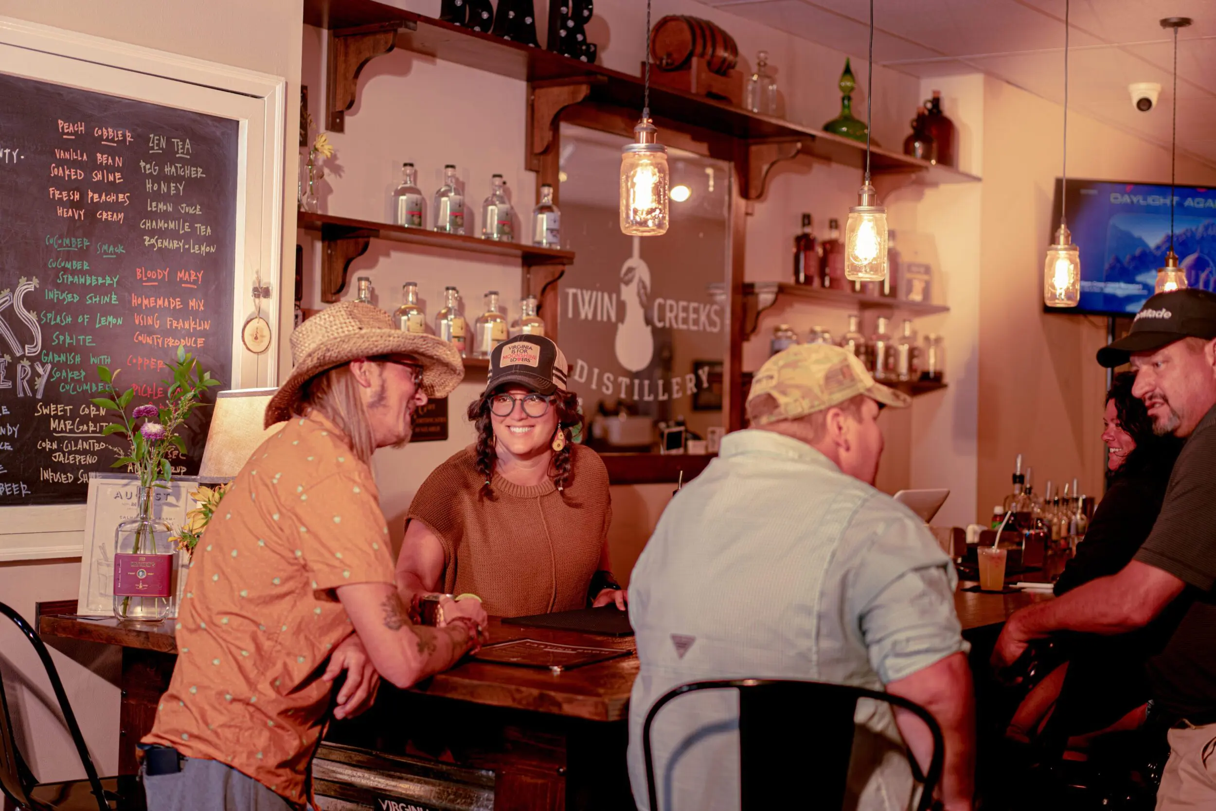 Two white men, one with a cowboy hat and one with a baseball cap, sit at a the bar at Twin Creeks Distillery in Franklin County, VA. The man in the cowboy hat is talking happily to the female bartender, who's facing toward him with a big smile on her face.