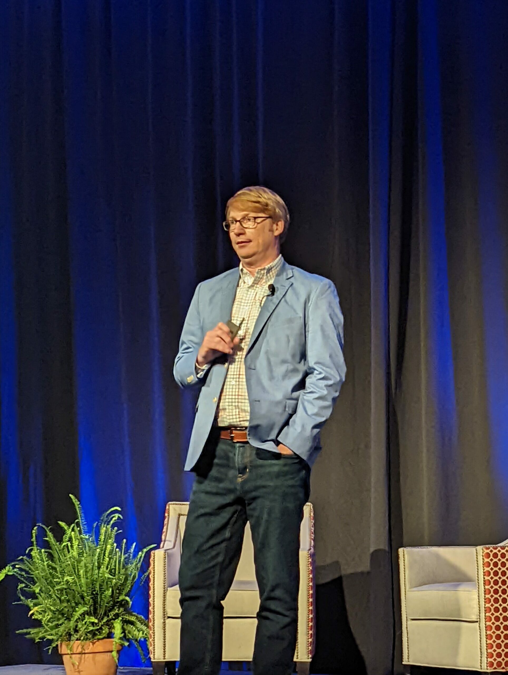 White man with blue blazer stands on stage to give presentation