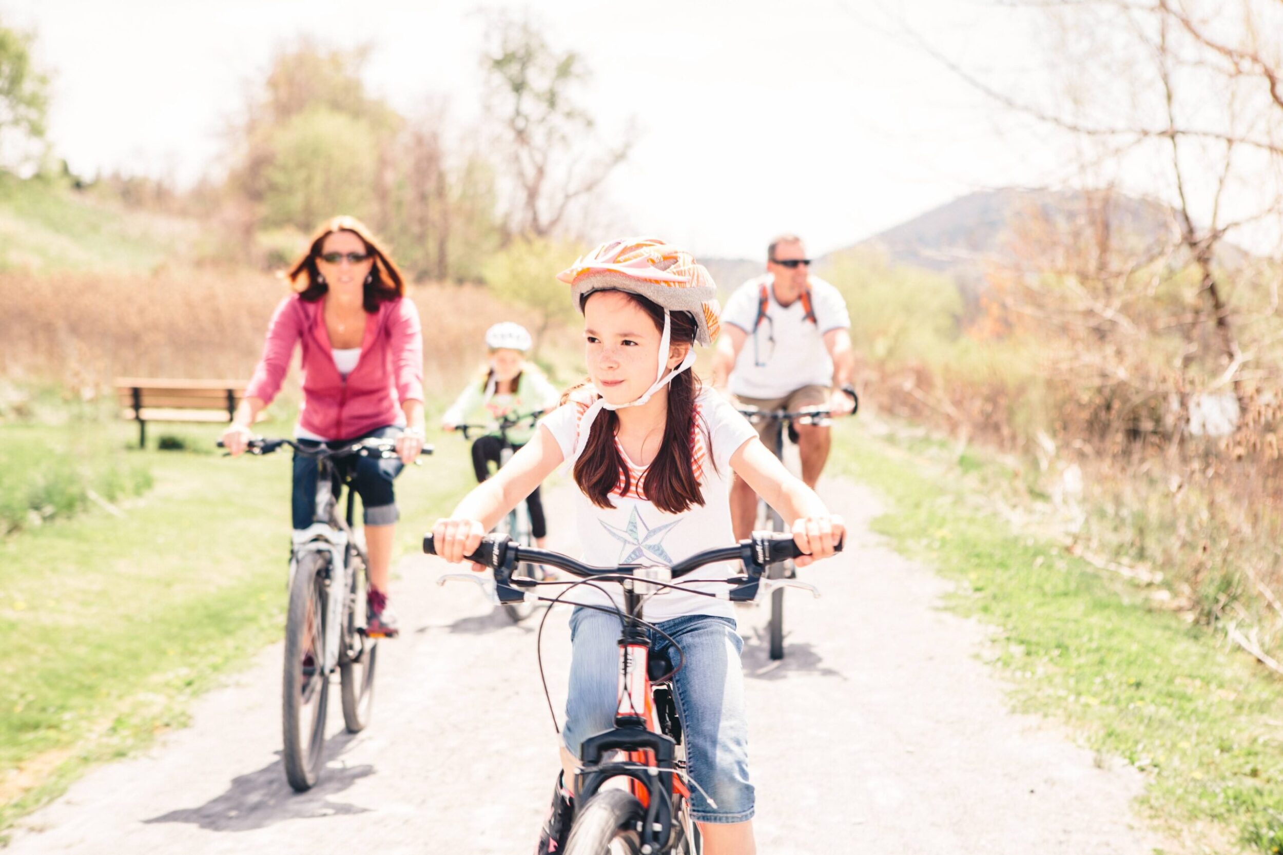 Young girl wearing a helmet bikes on a gravel path in Botetourt County, VA. Her mother, father, and younger sibling bike behind her in the background. It's a sunny day in early spring at the trees are just starting to bud.