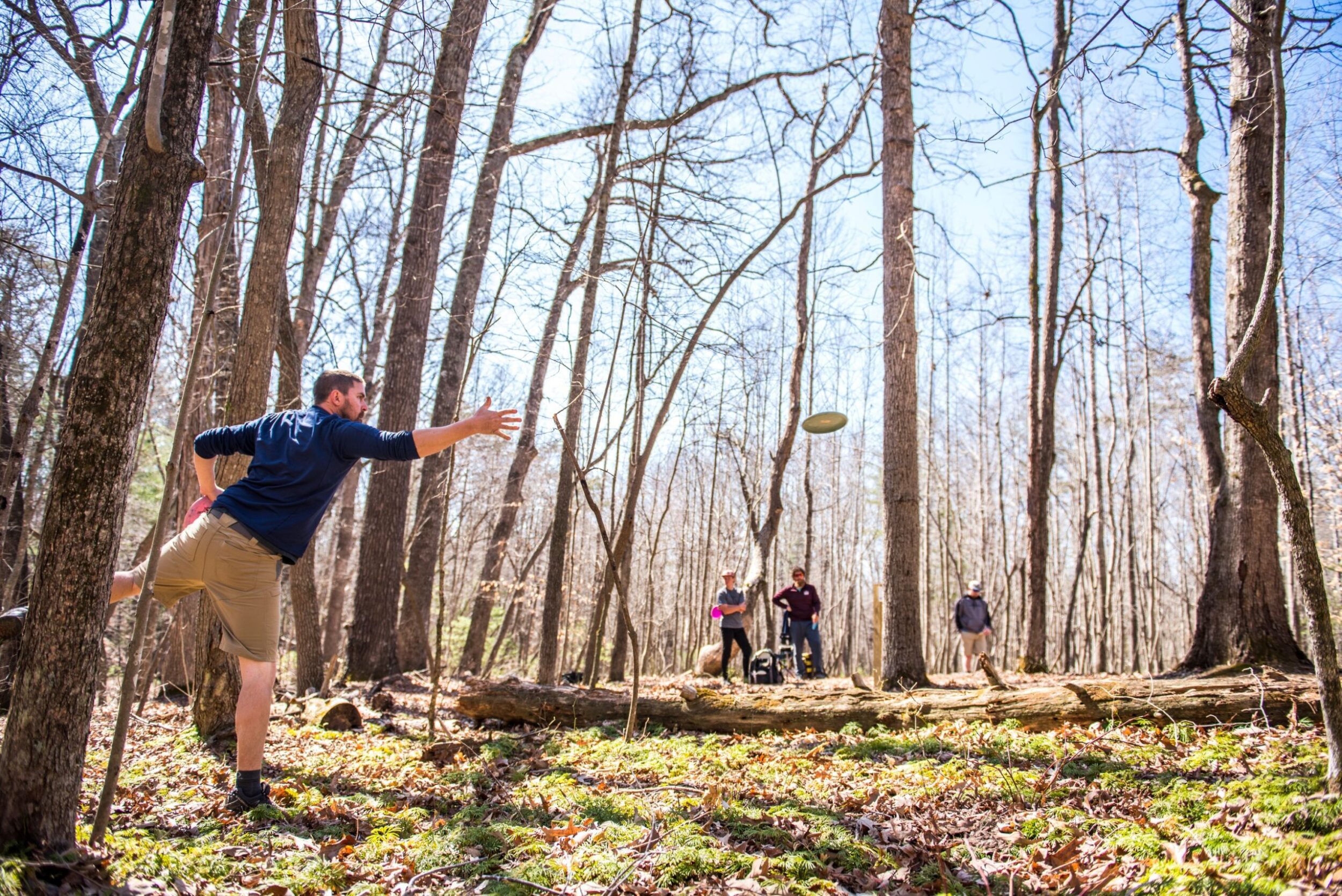 White man throws a disc golf frisbee in Waid Park on an early spring day. Three other white men in his party look on in the background as he throws.