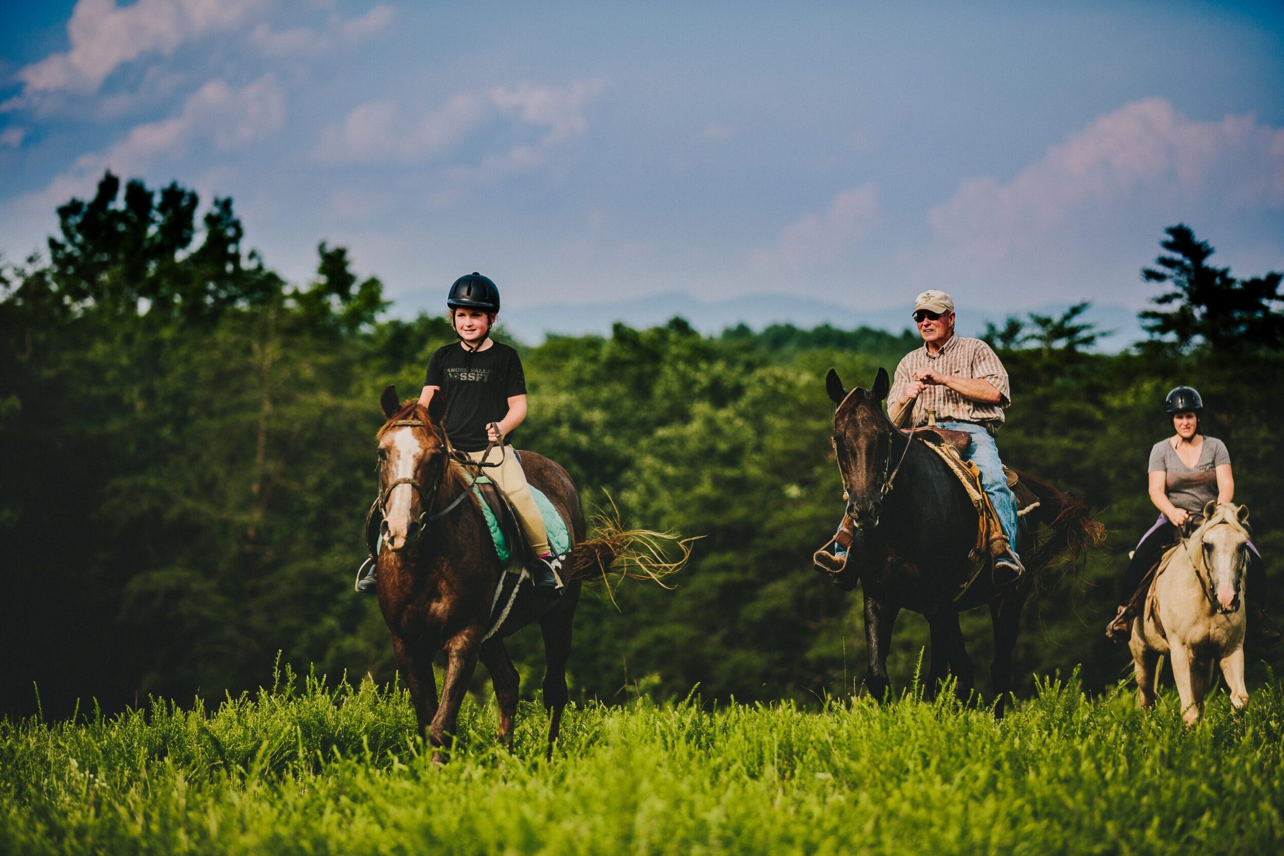 A young girl wearing a black helmet rides on a horse in a Botetourt County. Two adults ride horses in the background. It's a summer day during golden hour with tree-covered mountains in the background.