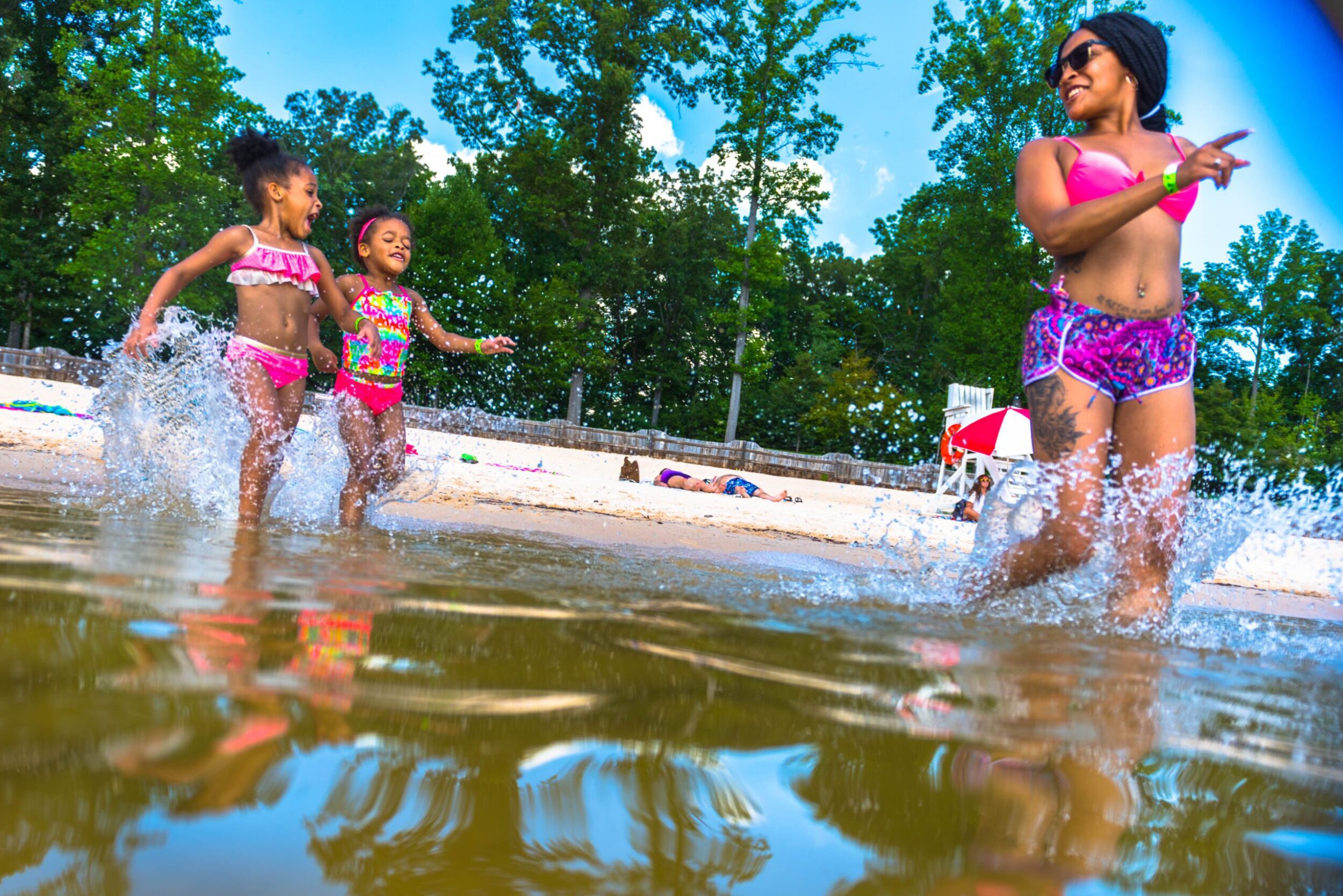 A smiling, Black mother in a bikini runs into the water with her two daughters, smiling and in swim suits, chasing after her.