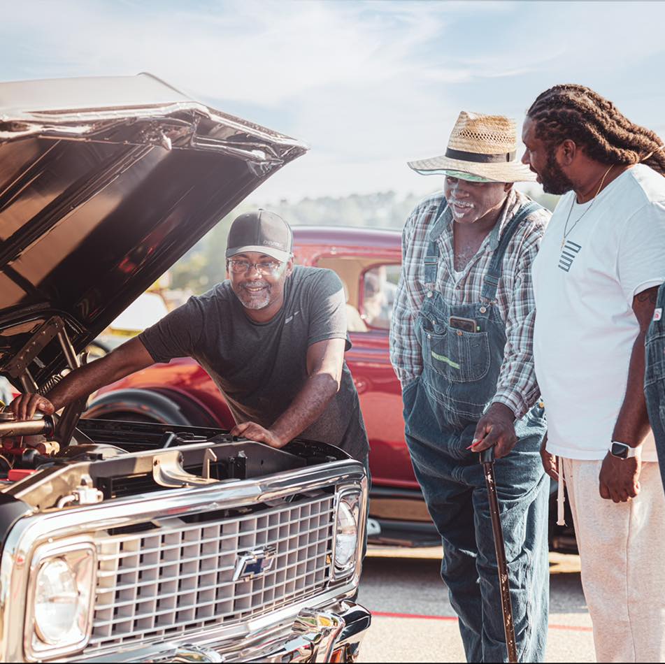 A Black man wearing a baseball cap is under the hood of a classic car and appears to be fixing something. Two other Black men watch him and smile.