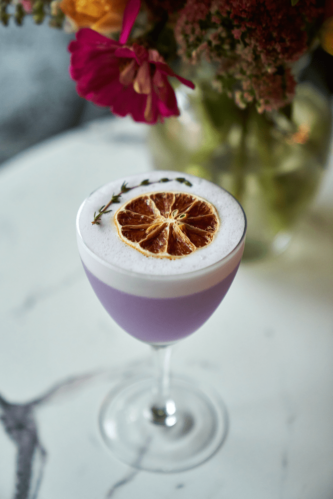 Fancy purple cocktail in a wine glass on a table with white table cloth and flowers.