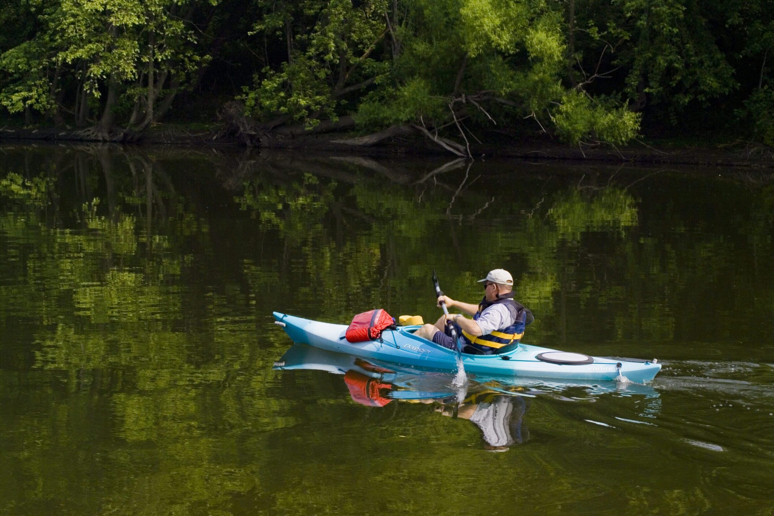 Older white man paddles along the James River in a blue kayak. There's lush greenery along the river on an overcast day in Botetourt County.