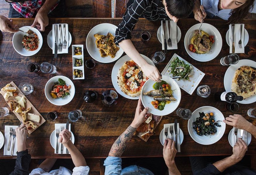 Overhead shot of a wooden table filled with plates of Italian food (fish, pasta, bread, oils) with arms extending across to grab plates.