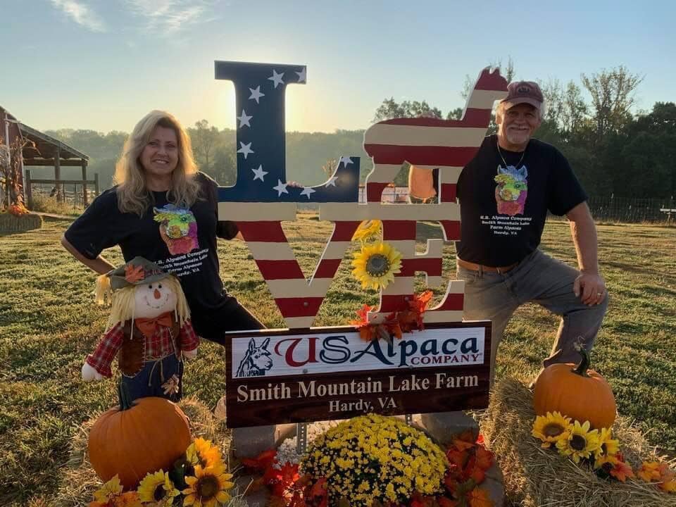 Two people pose in front of a "LOVE" sign that is decorated with fall decor. The sign reads "U.S. Alpaca Smith Mountain Lake Farm"