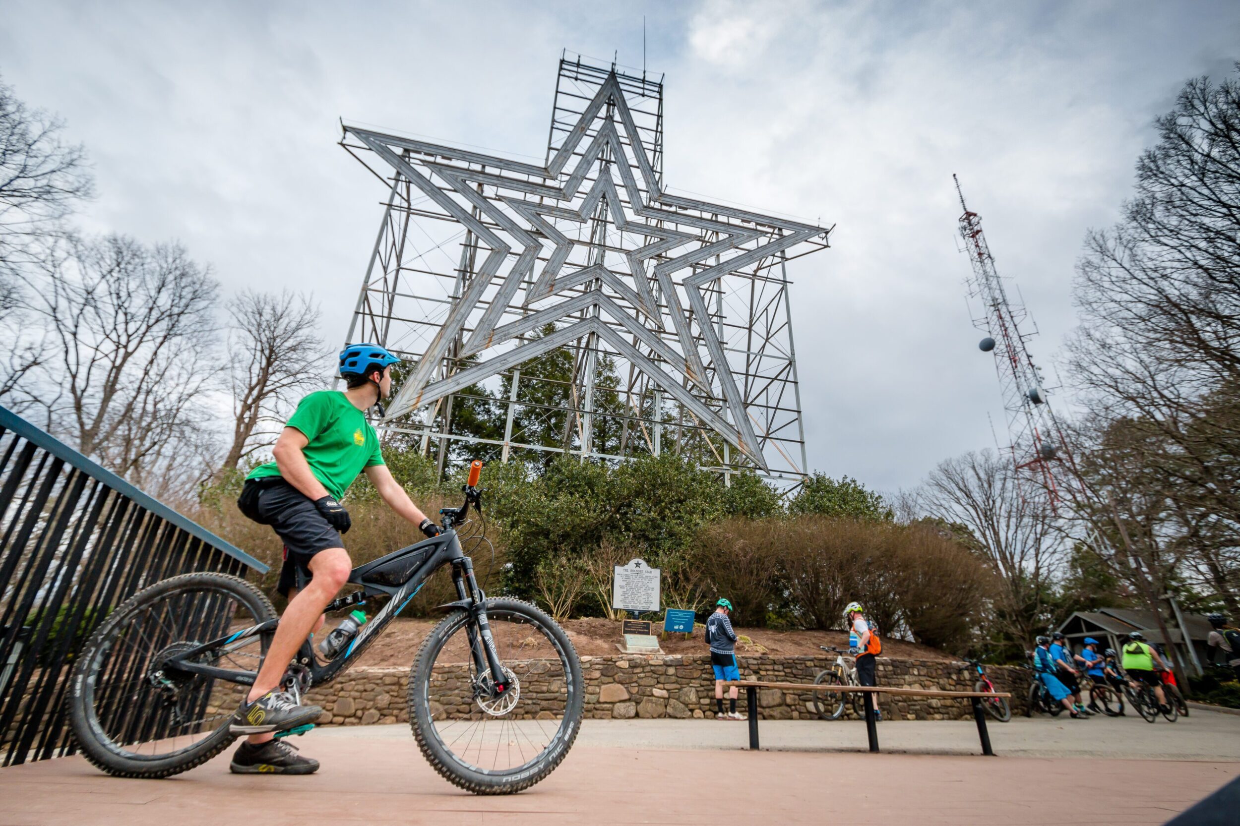 Mountain biker sits on bike and looks back at the Mill Mountain Star atop Mill Mountain. There are other cyclists in the background riding away from the overlook.