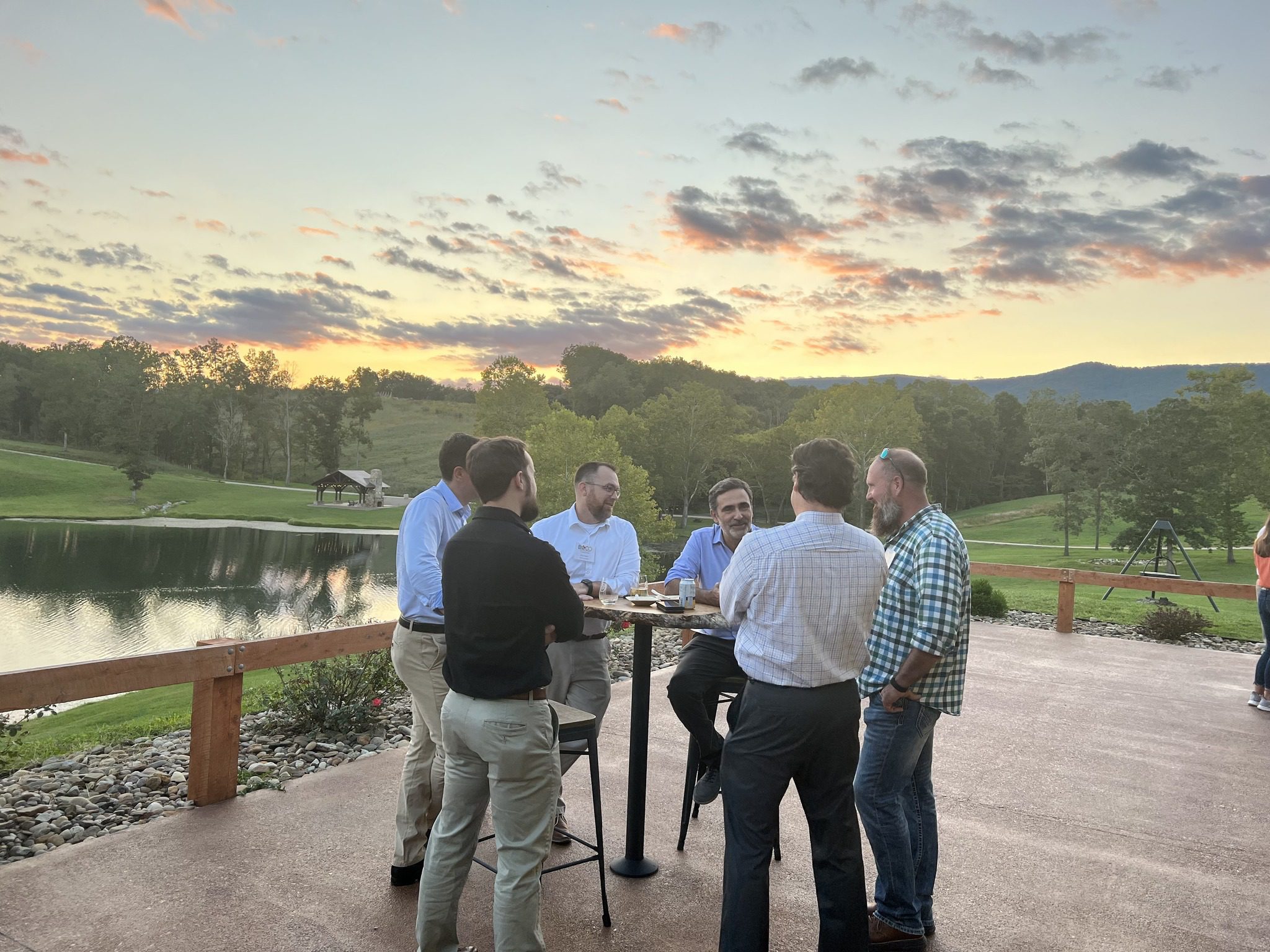 A group of men in business casual clothes at a cocktail table at the Preserve at Crooked Run with a beautiful sunset in the background.