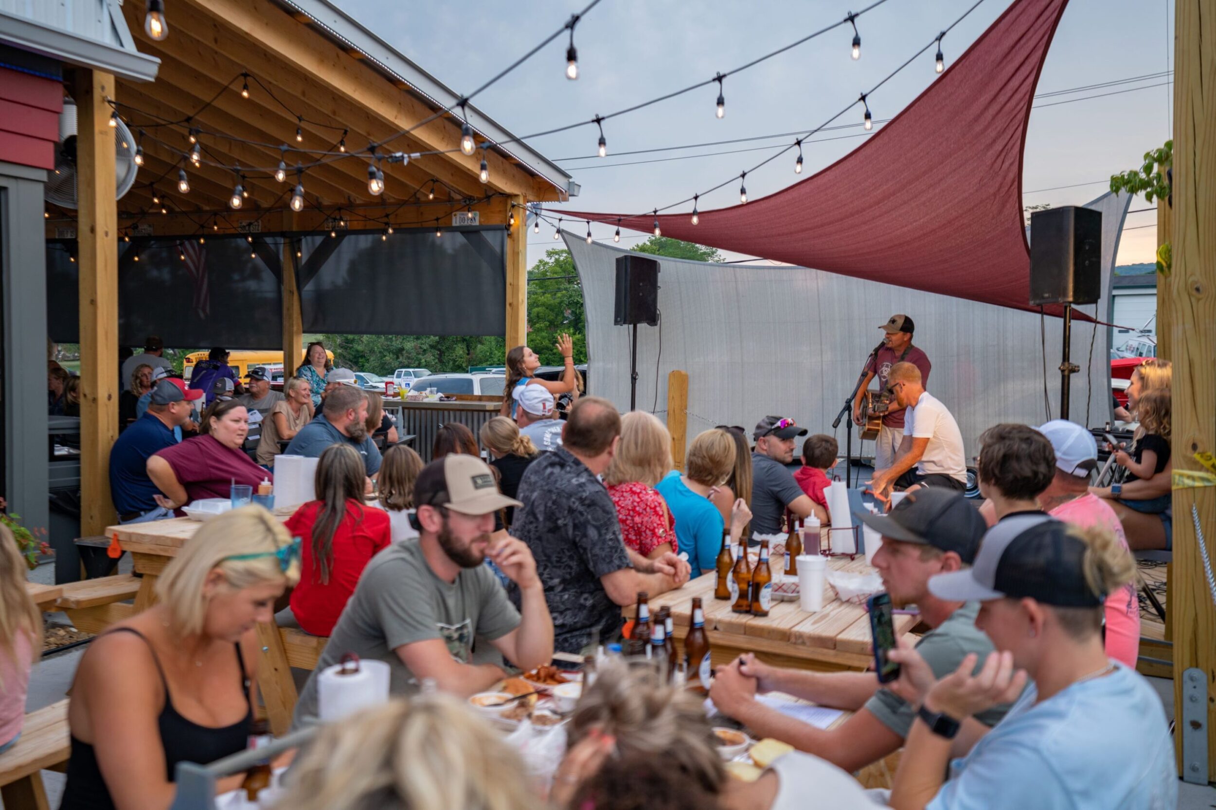 A crowd of people are seated on the outdoor patio of Smokehouse in Rocky Mount. A solo musician is seated on a stage performing for the diners.