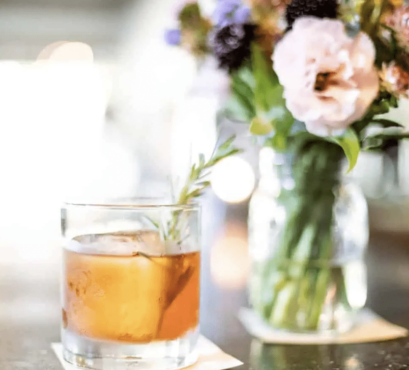 Fancy cocktail in a whiskey glass next to wild flowers in a vase atop a dark wooden table.