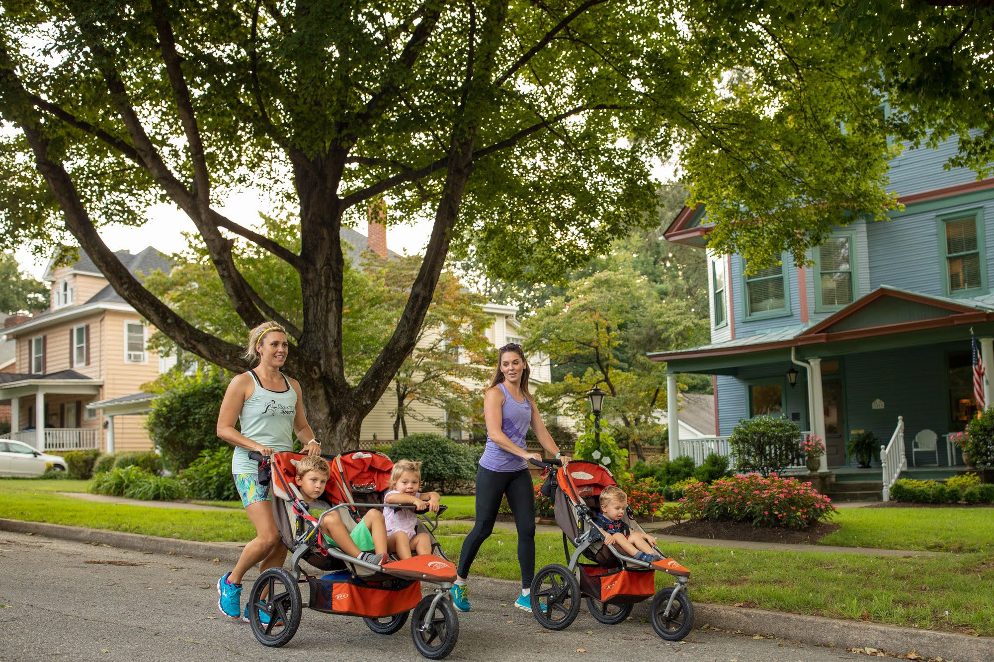 Two white women in athletic gear walk push their children in strollers with beautiful Victorian and Colonial homes in the background