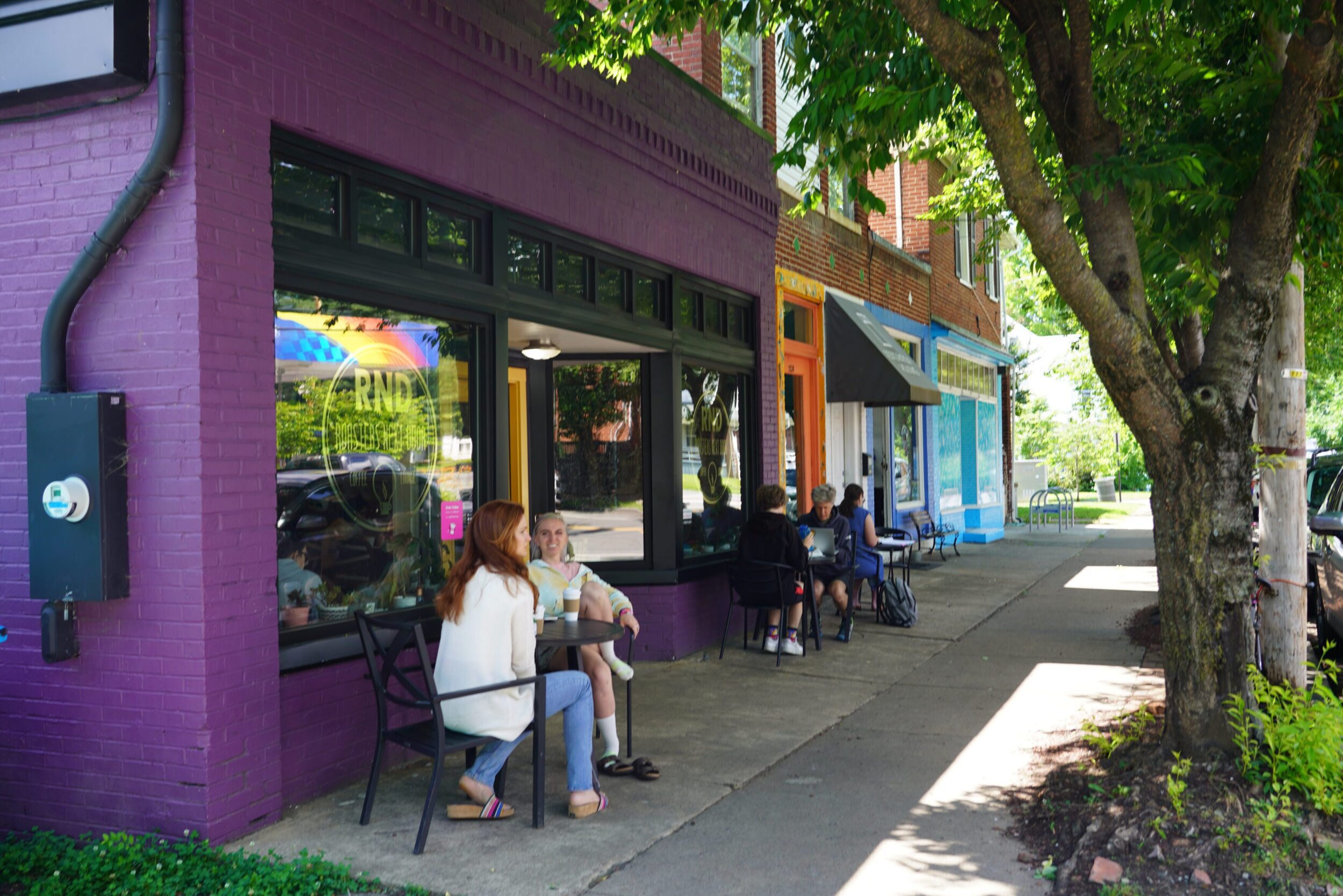 A line of colorful storefronts along Main Street in the Wasena neighborhood of Roanoke, VA. There are two women sitting outside of a coffee shop and more people sitting at an additional table in the background.