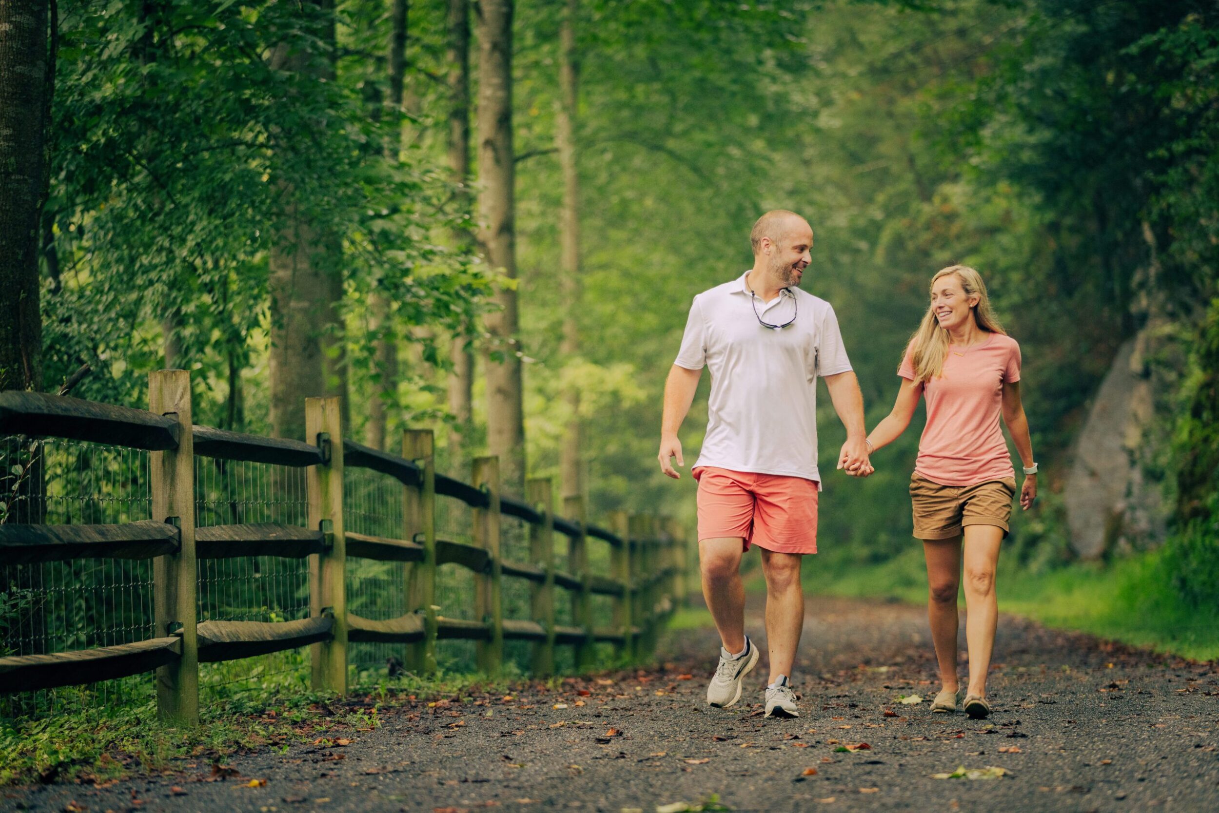 A white man and woman are holding hands and smiling as they walk down a gravel greenway path in the woods.