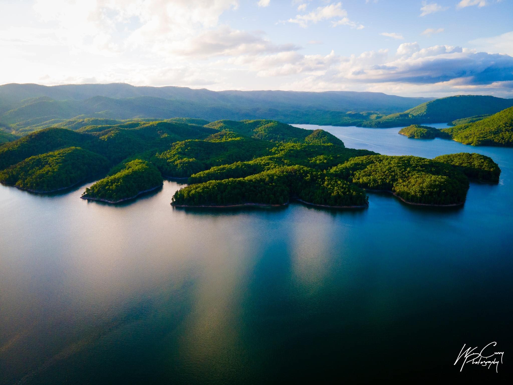 Aerial shot of Lake Moomaw on a beautiful summer day during golden hour. The lake is lined with mountains with green lumpy peninsulas in the water.