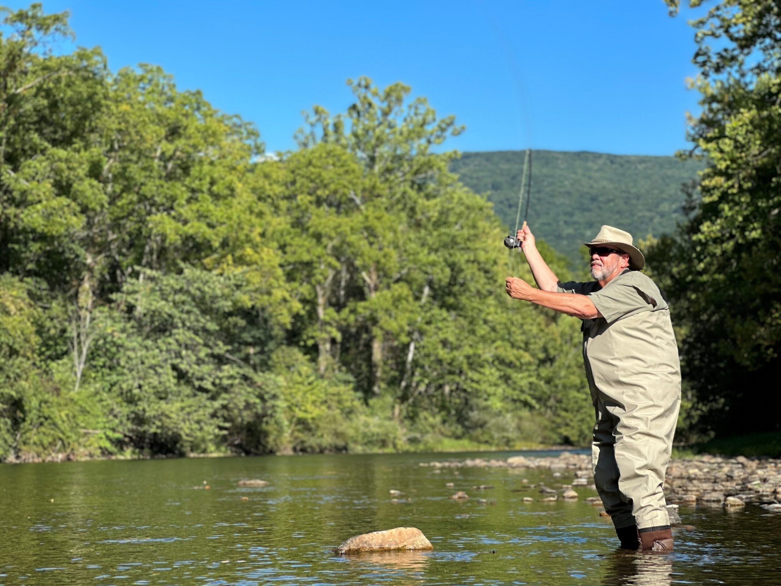 Older white man fly fishing in the river. The river is lined with green trees and mountains.