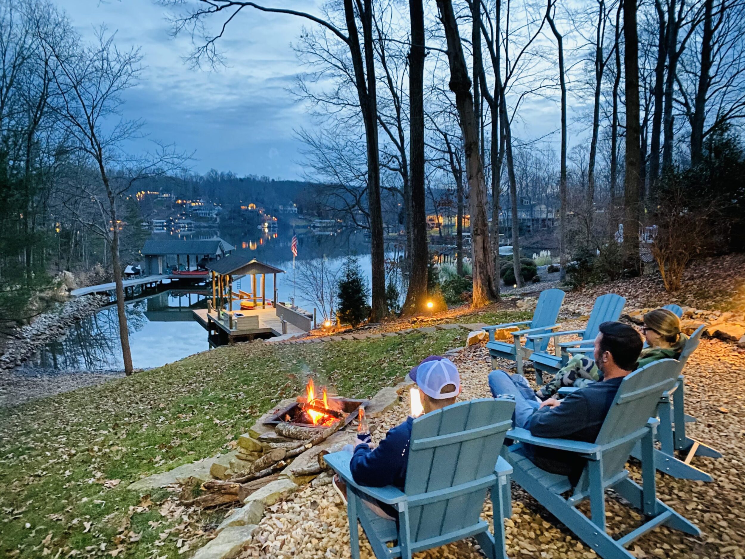 Three adults sit in adirondak chairs in front of a fire pit. They're on facing a dock in Smith Mountain Lake at dusk.