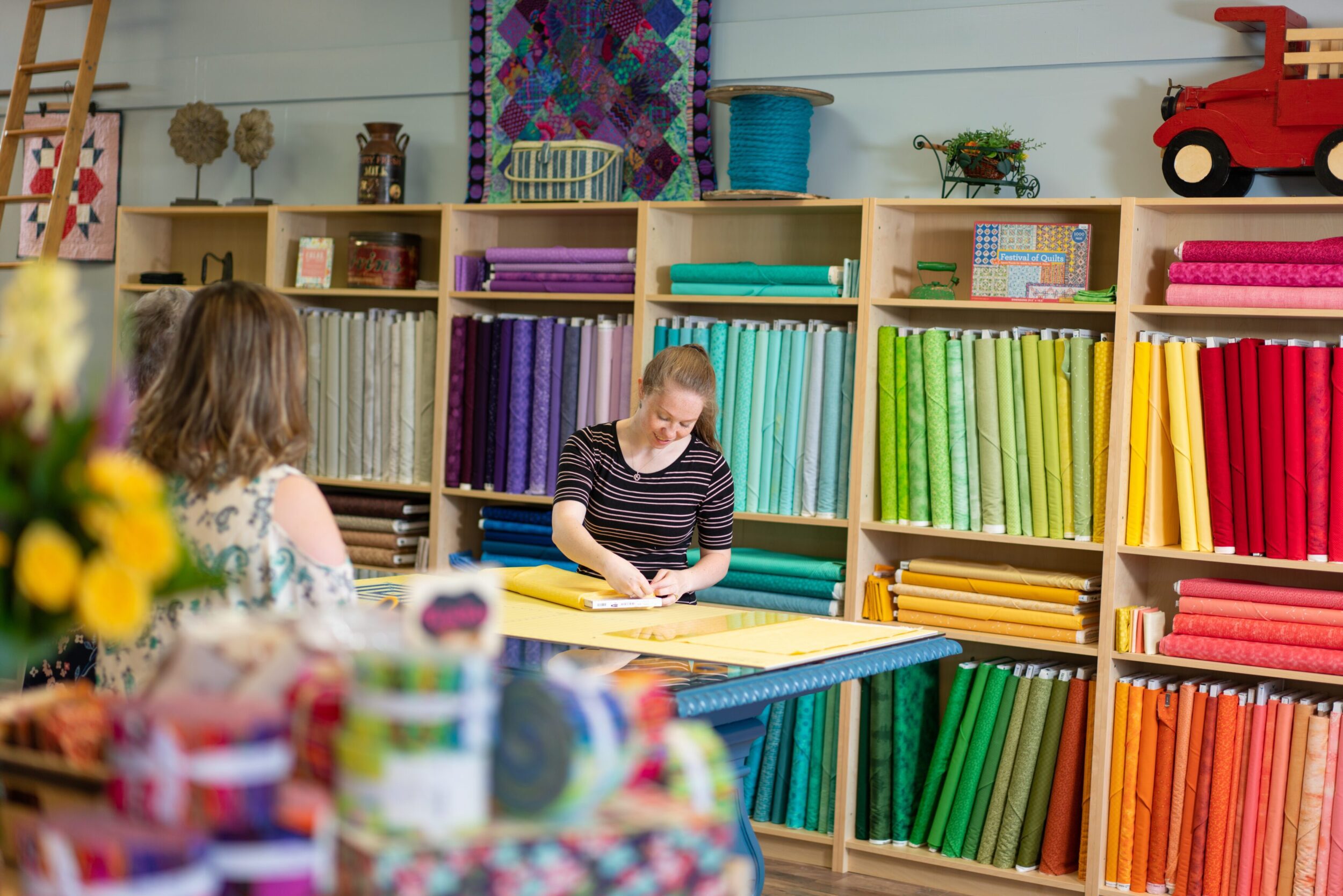 A young white woman measures out fabric on a table. Behind her is a rainbow of colorful quilting material lining the walls of the establishment.