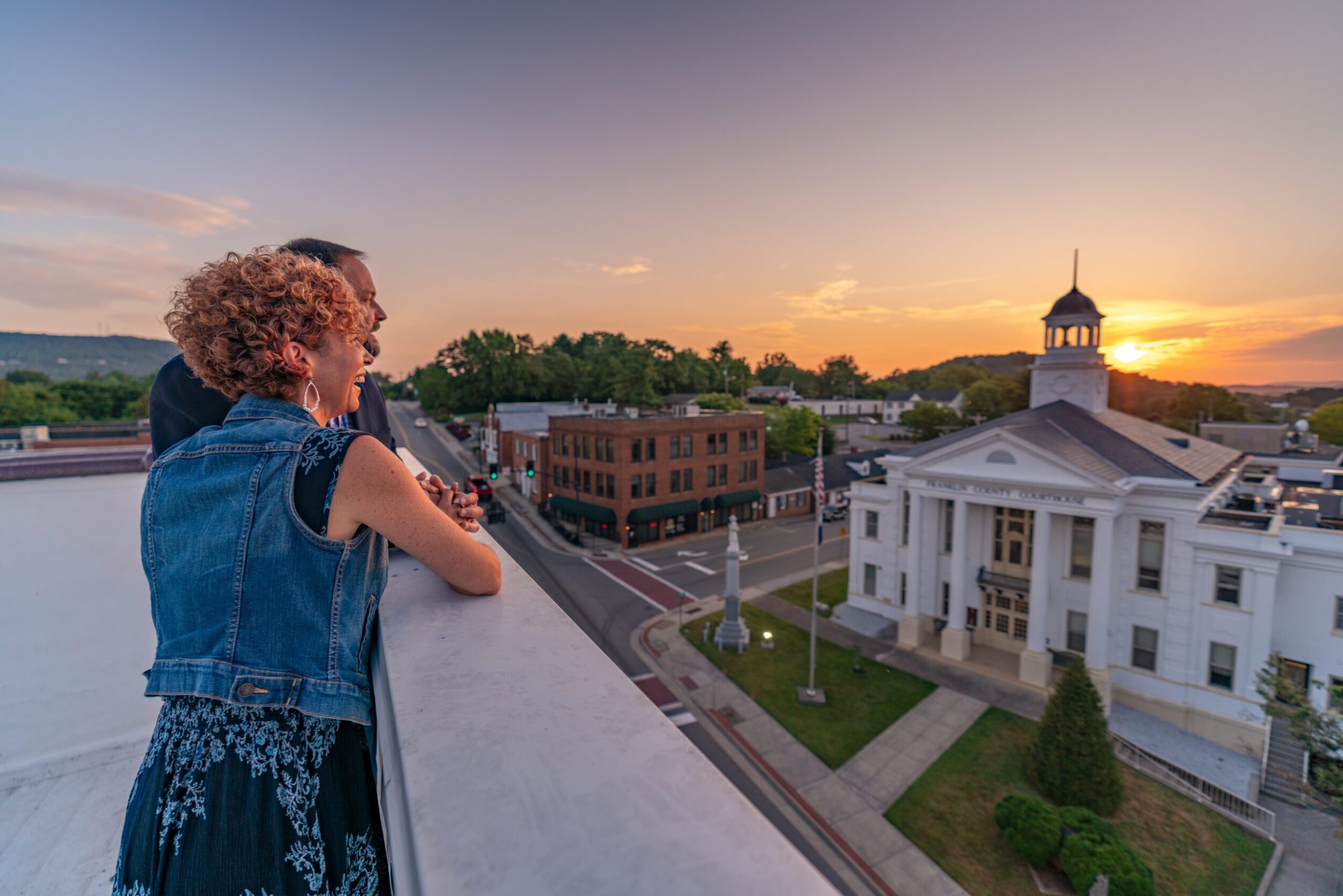 A middle-aged white couple stands on a roof-top overlooking downtown Rocky Mount at sunset on a summer evening.