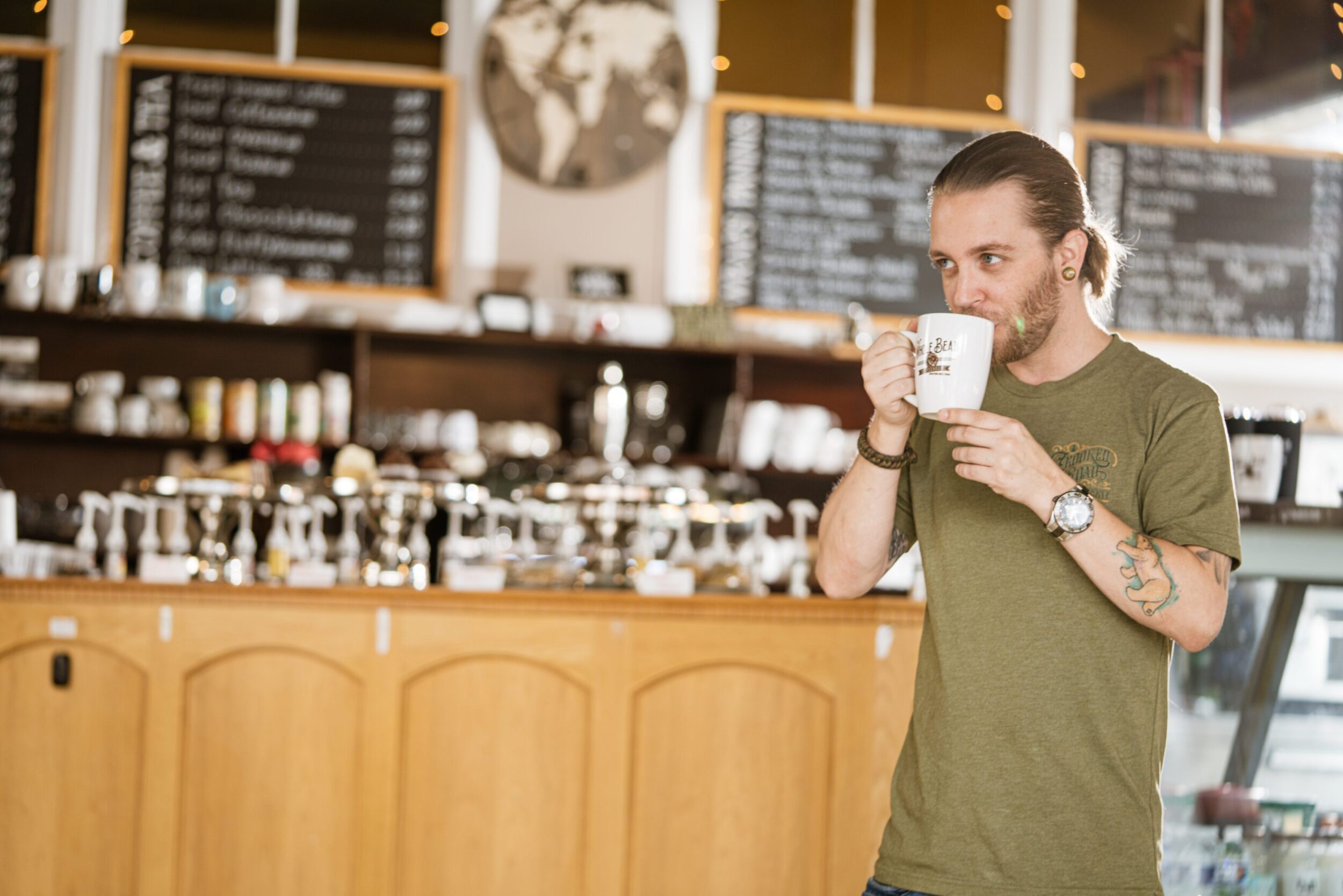 A man in his late twenties with a beard and ponytale sips from a white coffee mug in a trendy coffee shop in Rocky Mount, VA.