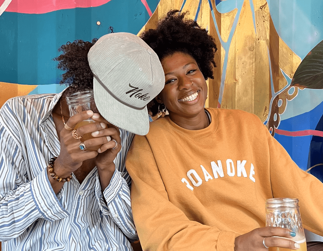 A Black man wearing a Noke hate covers his face as he laughs, and a young Black woman wearing a mustard yellow sweatshirt with "ROANOKE" written on it smiles at the camera. She is holding a beer from Golden Cactus Brewing, and there is a beautiful, colorful mural behind them.